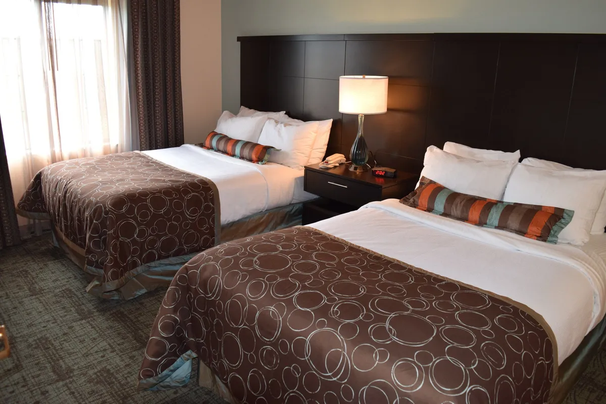 A comfortable interior with two double beds, a stylish dark headboard, decorative pillows, and a soft lamp. The room enjoys ample natural light, suggesting a welcoming and restful environment for families, friends, or colleagues.