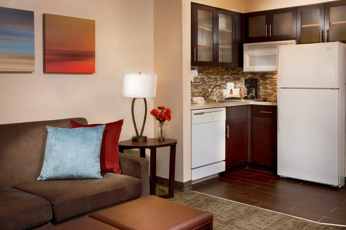 A spacious, open-plan living area featuring a comfortable sofa with decorative pillows, a nearby kitchen area, wall art, and a table lamp. The setting exudes a cozy and welcoming atmosphere, ideal for spending quality time together, relaxing, or entertaining. The sofa appears to be a pull-out, offering flexible sleeping arrangements for guests or larger groups.