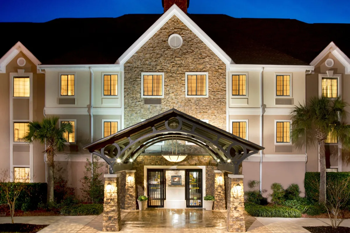This exterior image captures the property's welcoming entrance with an inviting awning, stone facade, and warmly lit windows. Evening light suggests a comfortable arrival no matter the hour, and the visible social activity hints at a vibrant, connected atmosphere.