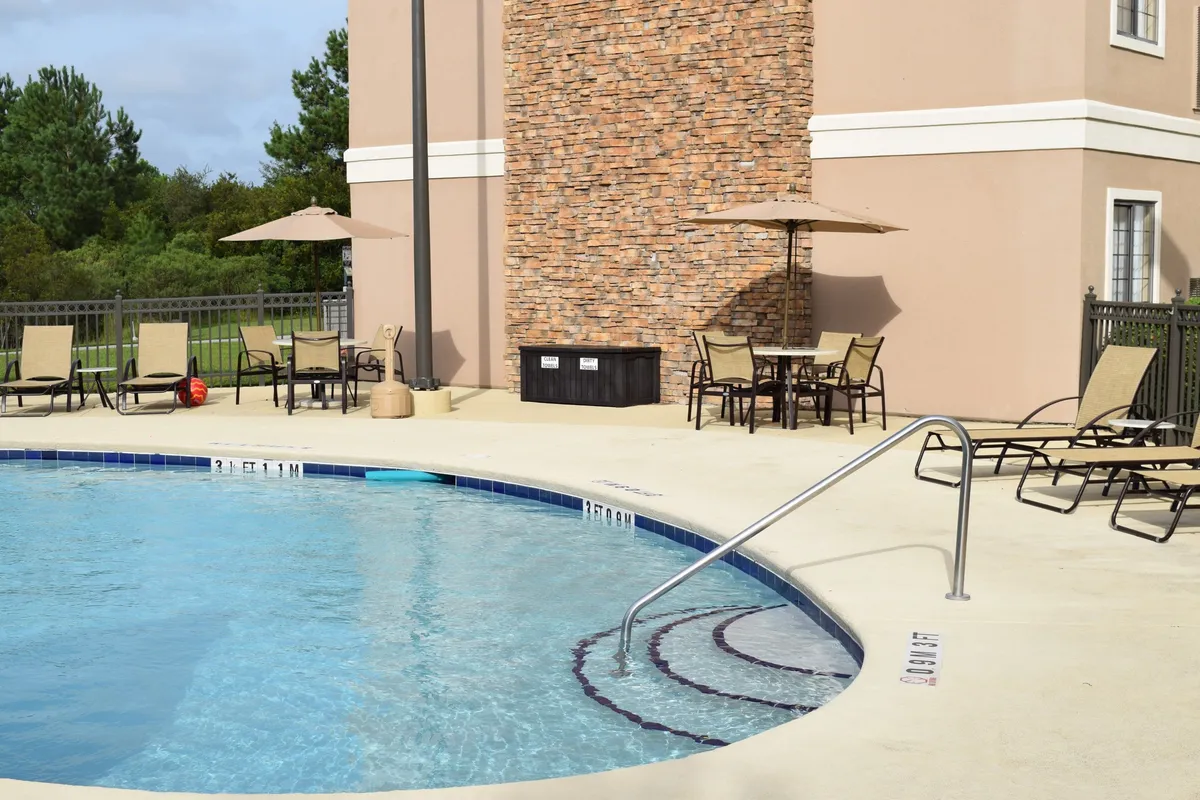 A sun-kissed outdoor pool surrounded by comfortable lounge chairs and shaded by umbrellas, with a stone wall adding to the inviting ambiance. Guests can be seen swimming, sunbathing, and socializing, creating a lively yet relaxed atmosphere.