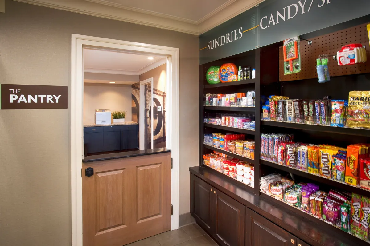 This image captures a welcoming interior scene featuring a well-stocked snack shelf, a sign reading 'THE PANTRY,' and a view toward an adjacent area—suggesting access to amenities beyond the suite itself. The casual arrangement and visible snacks evoke communal, hotel-like perks within a residential setting.